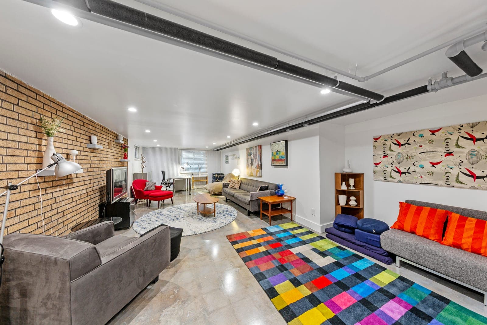 Long lower-level room with a blonde brick wall, color-block rug, and multiple seating areas