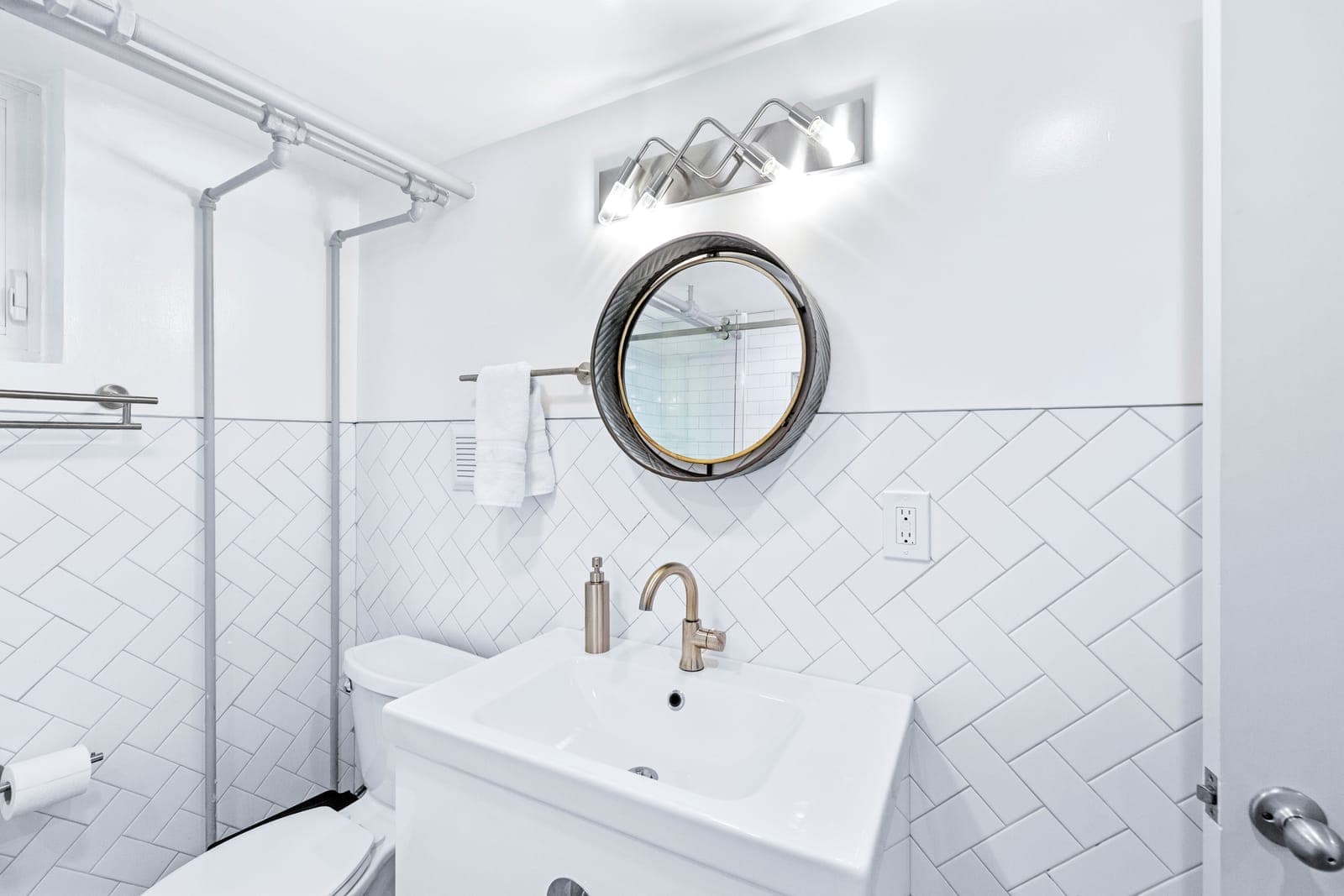 Hall bath vanity with a round framed mirror, sculptural sconce, and herringbone tile wainscot